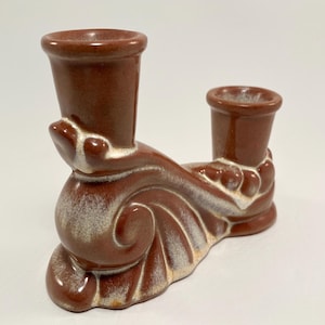 May include: A brown ceramic candle holder with a white glaze. The holder has two candle cups and is shaped like a stylized wave.
