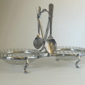 May include: A silver-toned serving dish with two clear glass bowls, each with a beaded rim. Two spoons hang from a central support. The dish has curved legs and is designed for serving condiments or small portions.