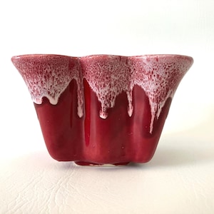 May include: A red ceramic planter with a unique, scalloped shape. The top edge features a white, dripped glaze effect, contrasting with the glossy red body. The planter has a low profile and a slightly textured surface.