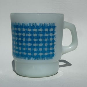 May include: White glass mug with a blue checkered pattern on the front. The mug has a rounded base and a curved handle. The blue pattern resembles a tablecloth design. The mug is likely vintage and suitable for hot beverages.