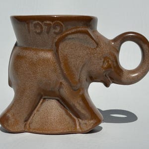May include: A brown ceramic mug shaped like an elephant, with the year "1979" embossed on the top. The mug has a rounded handle and a speckled finish, creating a vintage aesthetic. The elephant's trunk forms the spout.
