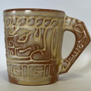 May include: A ceramic mug with a brown and tan glaze. The mug features a textured design with repeating patterns and a handle with a unique shape. The mug is suitable for hot or cold beverages.