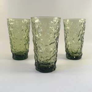 May include: Three vintage green textured drinking glasses. The glasses have a tall, cylindrical shape with a textured surface. The base of each glass is a darker shade of green. The glasses are arranged on a white surface.