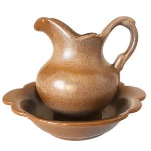 May include: A brown ceramic pitcher with a curved handle and spout, resting on a matching scalloped-edge bowl. The pitcher and bowl have a speckled, glazed finish, suggesting a rustic or vintage aesthetic.