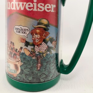 May include: Green and white Budweiser beer mug with a cartoon leprechaun holding a beer bottle and saying "This Bud's for you". The mug has a green handle and the words "Budweiser" and "Sh! Here!" printed on it.