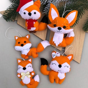 Puede incluir: Cinco adornos de zorro de fieltro naranja con detalles blancos. Los zorros tienen diferentes expresiones y algunos tienen lazos o bufandas. Los adornos cuelgan de una cuerda.