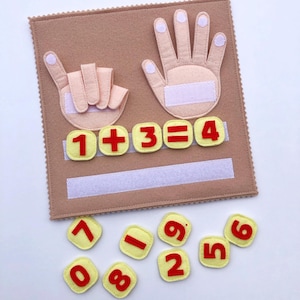Könnte beinhalten: Ein Filzbrett mit zwei Filzhänden, eine mit einem Finger nach oben und die andere mit allen Fingern nach oben. Das Brett hat einen Filzstreifen mit der Gleichung "1 + 3 = 4" darauf geschrieben. Es gibt auch Filznummern 0, 1, 2, 5, 6, 7, 8 und 9, die um das Brett herum verstreut sind.