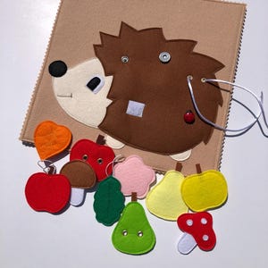 Peut inclure: Un jouet en feutre beige en forme de hérisson avec des épines marron, un visage blanc et un œil noir. Le jouet comprend divers fruits et légumes en feutre rouge, vert, jaune et orange. Une petite coccinelle rouge est attachée.