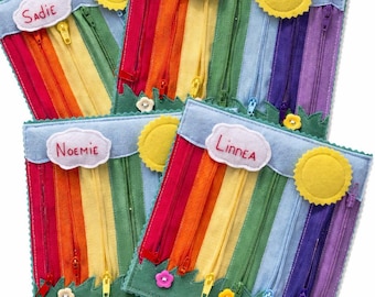 Puede incluir: Cuatro bolsas de fieltro con cremallera, cada una con un diseño de arcoíris, un sol amarillo y un nombre en una nube. Las bolsas tienen una base verde con detalles de flores. Los nombres en las bolsas son Sadie, Vanessa, Noemie y Linnea.