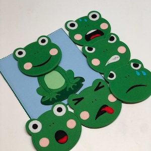 May include: A collection of green felt frog-shaped coasters with various facial expressions, including happy, sad, and surprised. A blue square coaster is also present, with a frog design. The coasters have pink cheeks and white eyes.