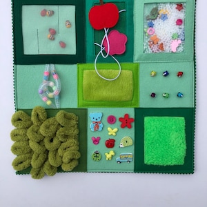 May include: A sensory activity board with a variety of textures and interactive elements. It includes a mesh window with beads, a felt apple and flower, a clear window with small shapes, and a section with buttons and a fuzzy texture. The board is predominantly green.