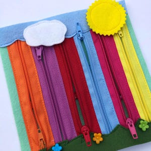 May include: A colorful felt activity board featuring a rainbow design with zippers in various colors. A yellow felt sun and a white cloud are attached to the board. Green felt grass with flower-shaped buttons and a blue sky complete the design.