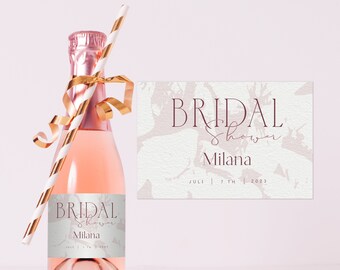 May include: A pink mini champagne bottle with a white label that says "Bridal Shower Milana July 7th 2023". The bottle is decorated with a gold ribbon and a striped straw. A white card with the same text is in the background.