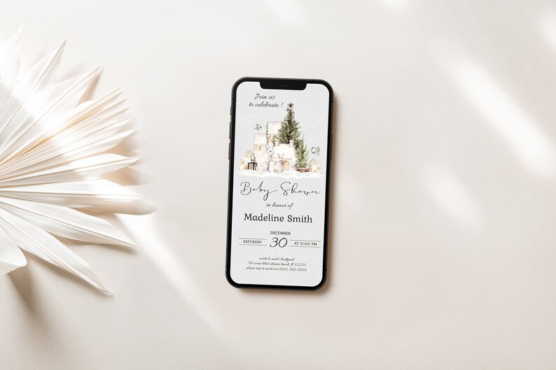 May include: A smartphone displaying a digital invitation for a baby shower. The invitation features a watercolor illustration of a winter scene with a Christmas tree, houses, and a lantern. The text reads "Baby Shower in honor of Madeline Smith, Saturday, December 30 at 1:00 PM".