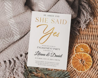 May include: White card with gold lettering announcing an engagement party for Liliana & Daniel. The card reads "He asked and she said Yes" with a gold script font. The date and time of the party are listed below, along with the address and RSVP information.