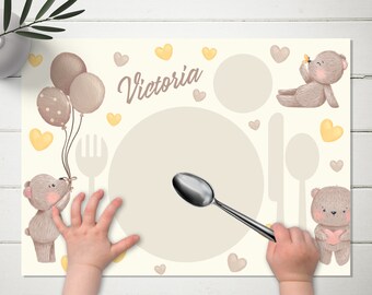 May include: A beige placemat with a brown bear holding balloons, a brown bear holding a heart, and a brown bear sitting with a butterfly. The placemat has a large circle in the center for a plate, and a fork and spoon are shown on the placemat. The placemat has the name "Victoria" written on it.