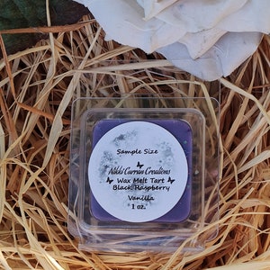 May include: A square, clear plastic container holding a purple wax melt tart. The label reads "Sample Size" and lists the scent as "Black Raspberry Vanilla." The container is nestled in straw, with a large, cream-colored flower in the background.