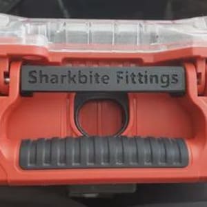 May include: A red plastic storage case featuring a black handle with a textured grip and a black label plate embossed with the text Sharkbite Fittings. The case includes a clear plastic lid section and a central circular opening for access.
