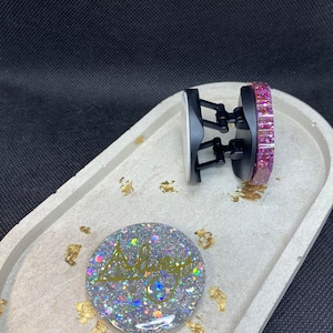 Peut inclure: Deux poignées de téléphone, l'une avec un motif de paillettes roses et l'autre avec un motif de paillettes argentées et le mot "Always" écrit en or. Les poignées sont sur un plateau gris avec des paillettes dorées.