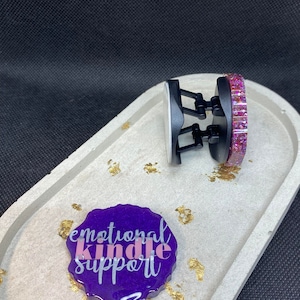 Peut inclure: Un porte-clés en résine violette avec le texte "emotional kindle support" en lettres blanches et roses. Le porte-clés est sur un plateau gris avec des paillettes dorées. Un support de téléphone noir avec un design de paillettes roses est également sur le plateau.