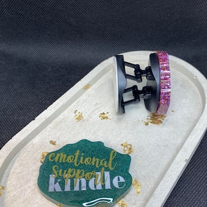 Peut inclure: Un dessous de verre en résine verte avec le texte "emotional support kindle" en lettres dorées. Un support de téléphone noir avec une base en paillettes roses est sur un plateau blanc.
