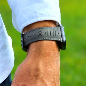 Könnte beinhalten: Graues Leder-Uhrenarmband mit Spotify-Logo und Musikwellenmuster. Das Armband ist an einer schwarzen Smartwatch befestigt. Das Armband wird am Handgelenk getragen, mit einem weißen Hemdärmel im Hintergrund.