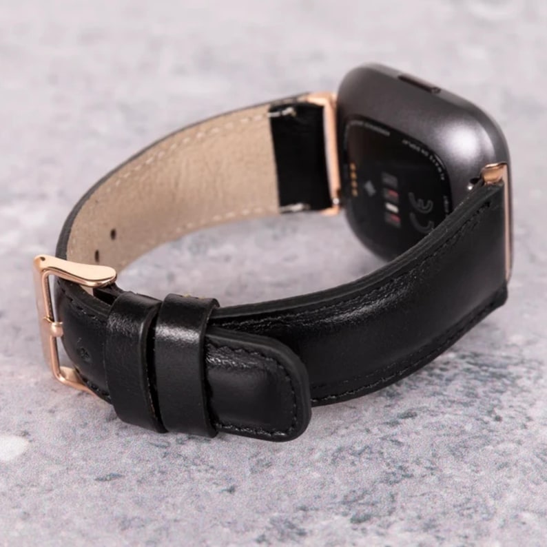 Peut inclure: Un bracelet de montre en cuir noir avec une boucle en or rose et une montre connect&eacute;e noire. Le bracelet de montre est con&ccedil;u pour un Fitbit Versa.