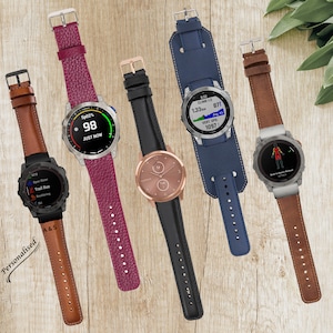 May include: Collection of smartwatches with various leather and silicone bands. The watches display different interfaces, including heart rate, workout data, and time. Band colors include brown, burgundy, black, and blue. The watches are displayed on a light wood surface.