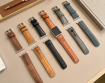 May include: A collection of leather watch bands in various colors including tan, black, gray, and blue, with one band attached to a silver smartwatch. Some bands feature custom engravings like the word faith, a paw print with the name tom, and a letter y.
