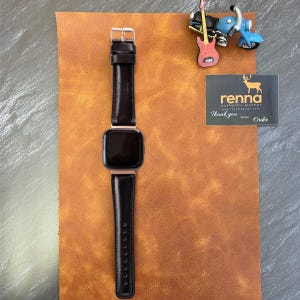 Peut inclure: Un bracelet de montre en cuir noir avec une boucle argent&eacute;e et un cadran noir. Le bracelet de montre est sur une surface en cuir marron. Le texte "renna authentic leather" est visible sur une carte blanche.