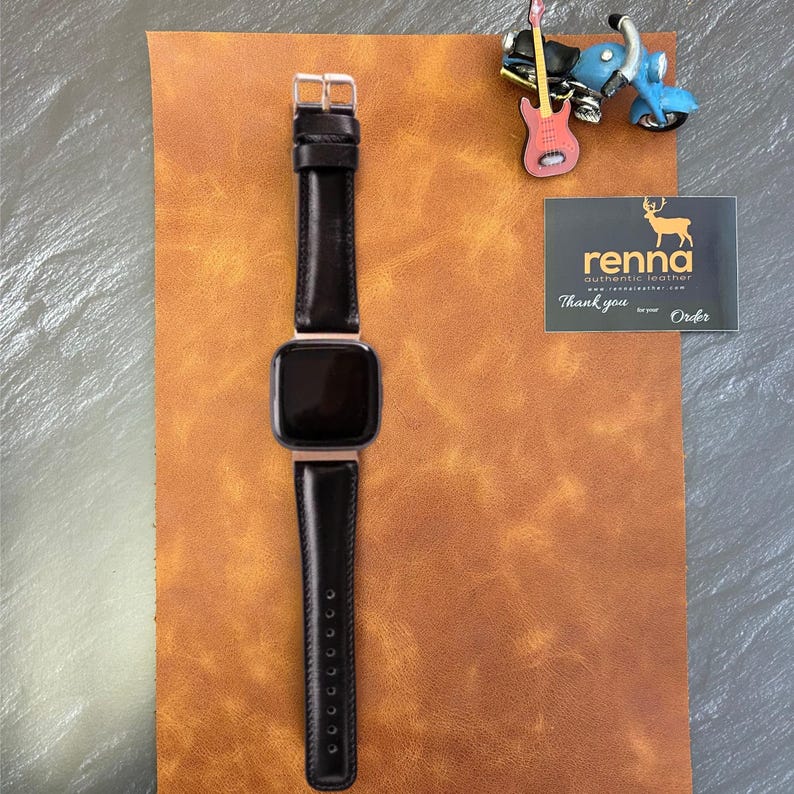 Peut inclure: Un bracelet de montre en cuir noir avec une boucle argent&eacute;e et un cadran noir. Le bracelet de montre est sur une surface en cuir marron. Le texte "renna authentic leather" est visible sur une carte blanche.