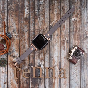 May include: A gray leather watch band with rose gold accents and a black square watch face. The watch is laying on a wooden surface with a vintage camera and the word "renna" spelled out in wooden letters.