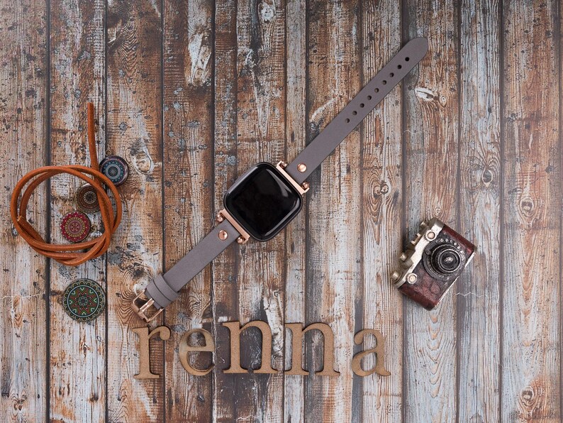 May include: A gray leather watch band with rose gold accents and a black square watch face. The watch is laying on a wooden surface with a vintage camera and the word "renna" spelled out in wooden letters.