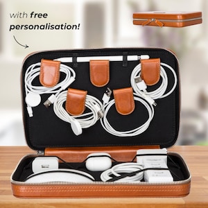 Puede incluir: Organizador tecnológico de cuero marrón abierto con interior negro, que contiene cables blancos, cargadores y un lápiz óptico. El organizador tiene sujetacables de cuero marrón y cierre de cremallera. El texto "with free personalisation!" es visible.