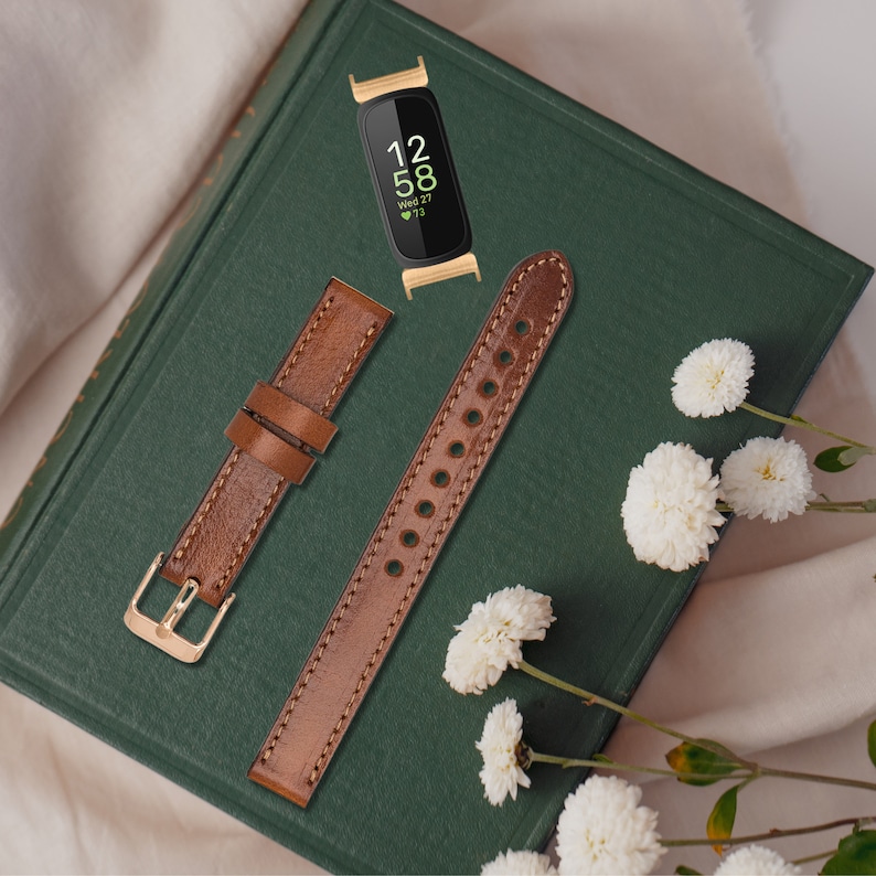 May include: Two brown leather watch bands for a smart watch. The watch band on the left has a gold buckle. The watch band on the right has a row of holes. The watch band on the left is laying flat. The watch band on the right is laying on its side. The watch bands are on a green book with white flowers.