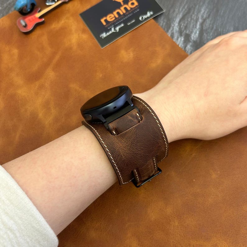 Puede incluir: Una correa de reloj de cuero marr&oacute;n con costuras blancas. La correa est&aacute; dise&ntilde;ada para adaptarse a un reloj inteligente con una esfera redonda. La correa se muestra en la mu&ntilde;eca de una persona.