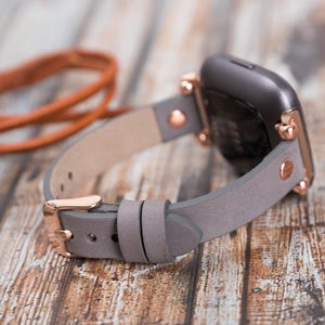 May include: A gray leather watch band with rose gold hardware. The band is shown attached to a smartwatch with a black face.