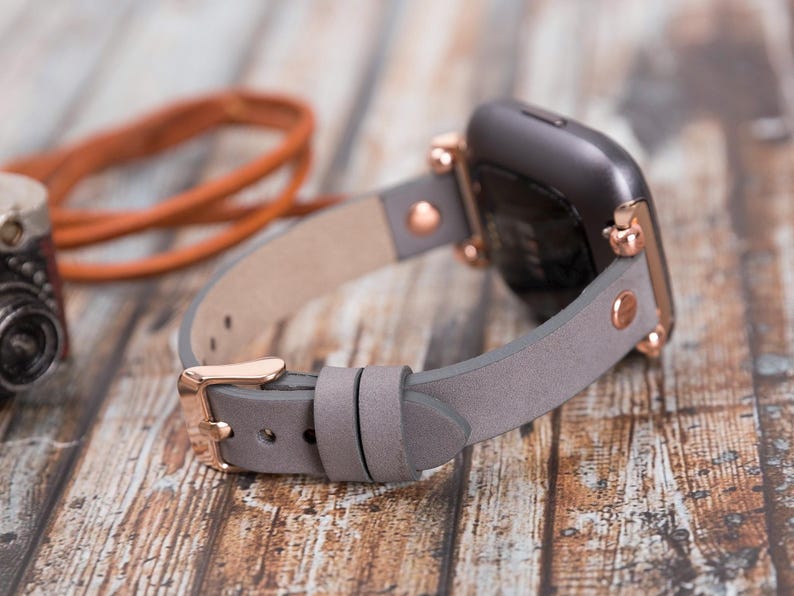 May include: A gray leather watch band with rose gold hardware. The band is shown attached to a smartwatch with a black face.