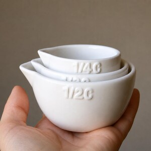 May include: Three white ceramic measuring cups, stacked together. The cups are marked with embossed measurements: 1/4 C, 1/3 C, and 1/2 C. Each cup has a pouring spout. The cups are held in a hand.