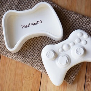 以下が含まれることがあります： 白いセラミック製のゲームコントローラー型の小物入れと蓋。蓋には茶色で「PapaLion100」の文字。コントローラーにはボタンと方向パッドがあります。ジュート布と木製の表面に置かれています。
