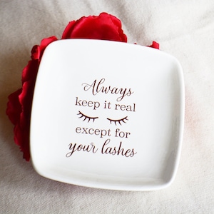 May include: White ceramic trinket dish with the text "Always keep it real except for your lashes" in brown lettering. The dish is decorated with a pair of closed eyes with eyelashes.