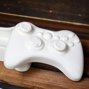 May include: White ceramic game controller shaped like a video game console controller. The controller has buttons and a raised area for the thumbsticks.