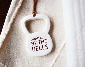 May include: A white ceramic kettlebell-shaped ornament featuring a brown leather cord at the top. The front surface displays the dark brown text "GRAB LIFE BY THE BELLS" in a clean, sans-serif font, centered within the smooth, rounded body.