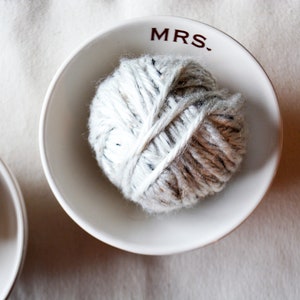 May include: Two white ceramic bowls with the words "MRS." and "MR." printed on them. Each bowl contains a ball of white and brown yarn.