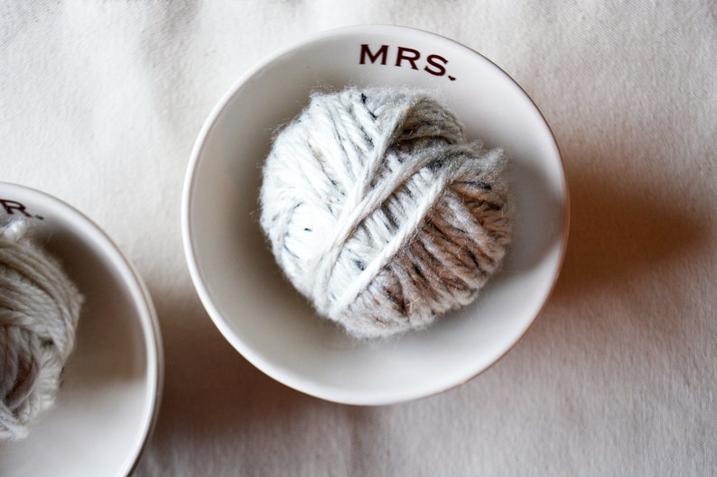 May include: Two white ceramic bowls with the words "MRS." and "MR." printed on them. Each bowl contains a ball of white and brown yarn.