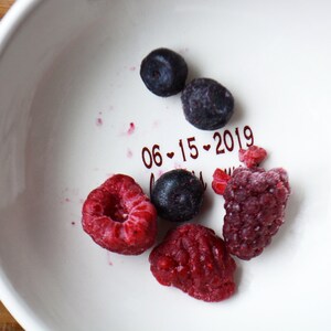 May include: A white ceramic bowl with a red inscription that reads "06-15-2019". The bowl contains a variety of frozen berries, including blueberries and raspberries.