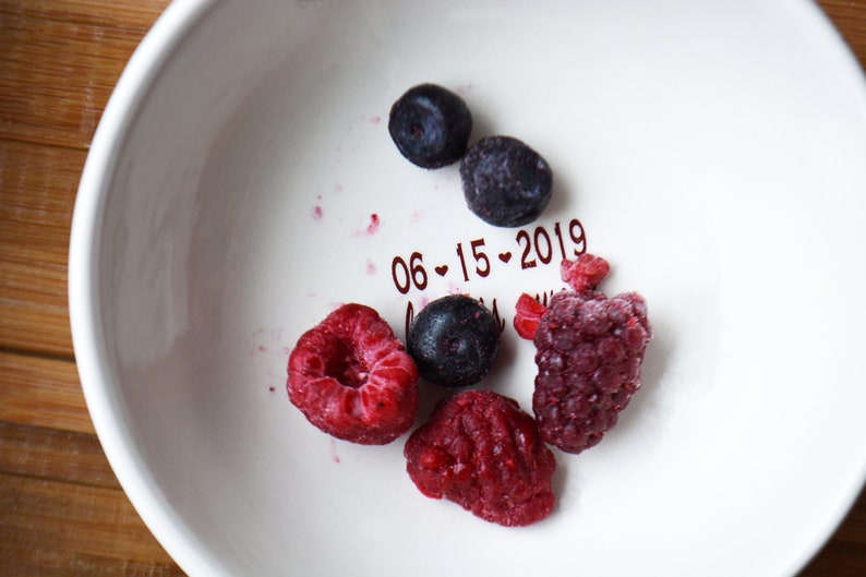 May include: A white ceramic bowl with a red inscription that reads "06-15-2019". The bowl contains a variety of frozen berries, including blueberries and raspberries.