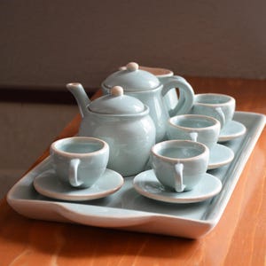 Puede incluir: Un juego de té de cerámica azul claro sobre una bandeja blanca. El juego incluye una tetera, un azucarero y cuatro tazas de té con platillos. El juego de té se exhibe sobre una superficie de madera.