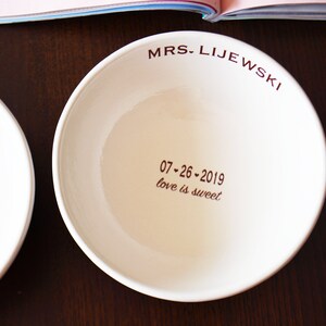 May include: Two white ceramic bowls with brown lettering. The bowl on the left says "SKI 2019 love is sweet". The bowl on the right says "MRS. LIJEWSKI 07-26-2019 love is sweet".