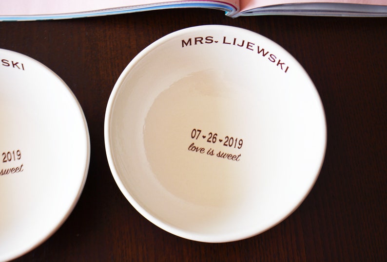 May include: Two white ceramic bowls with brown lettering. The bowl on the left says "SKI 2019 love is sweet". The bowl on the right says "MRS. LIJEWSKI 07-26-2019 love is sweet".